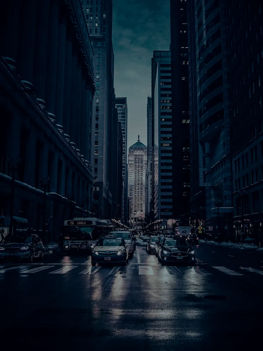 Avenida do centro financeiro ao entardecer, com torres de escritório dos dois lados e o asfalto molhado refletindo as luzes.